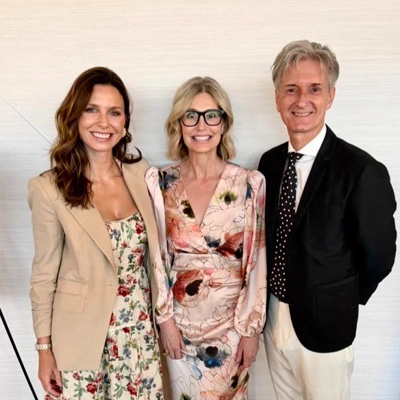 Three people smiling, standing side by side. The person on the left is wearing a floral dress and beige jacket. The person in the middle is in a floral dress. The person on the right is wearing a black suit with a polka dot tie.