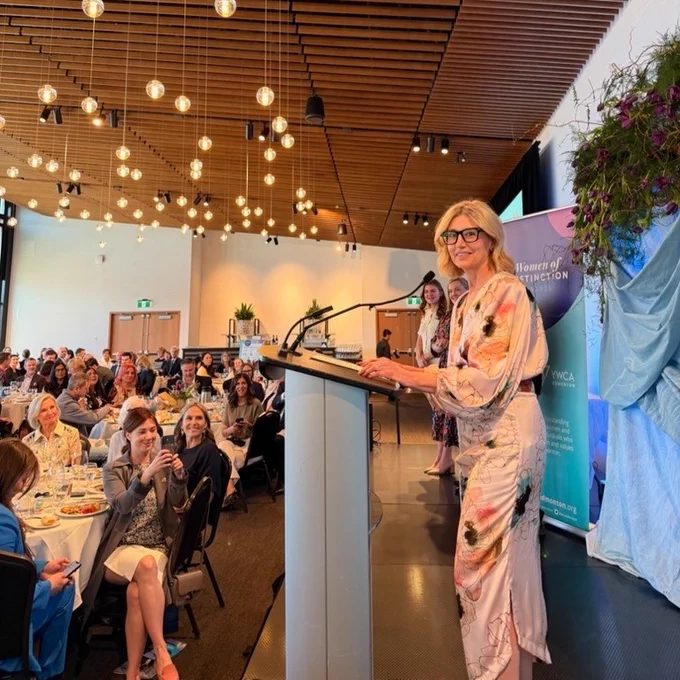 A person stands at a podium speaking to a seated audience during a formal event in a modern hall. The speaker wears a light-colored, floral-patterned outfit and glasses. Rows of attendees sit at tables with plates, glasses, and programs. The room features a wood-slat ceiling with many hanging globe lights, banners near the front, and floral arrangements by the podium.