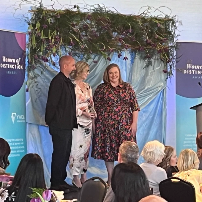 A group of three individuals stands on a stage under a floral arch. Two "Women of Distinction Awards" banners are visible in the background. Several people sit at tables in the foreground, watching the stage.
