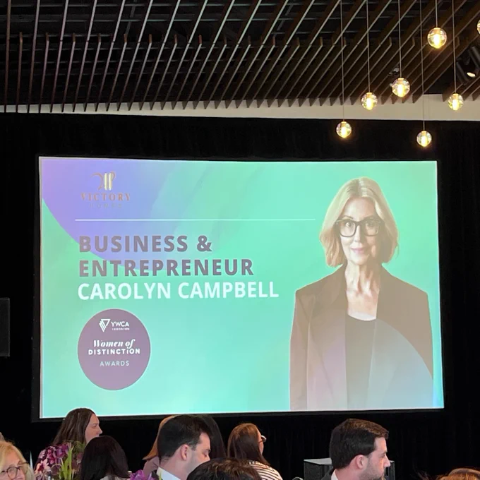 This image shows an event with a large screen displaying an award announcement. The screen features text: "Business & Entrepreneur Carolyn Campbell" and a badge for "YWCA Women of Distinction Awards." A photo of a person is on the screen. Several attendees are seated at tables in the foreground.
