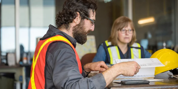 Two people at a table wearing reflective safety vests. One person is holding papers, and a yellow hard hat is on the table. A map is in the background.