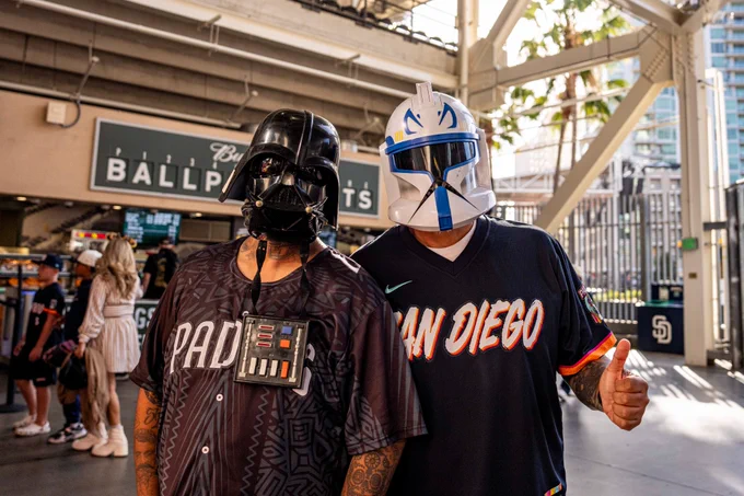 Photo of two Padres fans posing with Star Wars helmets on. The fan on the left is wearing a Darth Vader helmet, and the fan on the right is wearing a Captain Rex helmet.