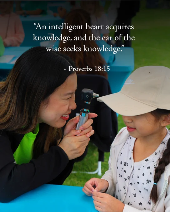 A cheerful woman holds an otoscope or ear examination device up to a young girl's face, demonstrating its purpose. They are outdoors in an educational setting. A blue table, green grass, and other event attendees are visible in the background and foreground. Above reads a quote from Proverbs 18:15.
