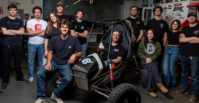 A group of individuals posing around an off-road vehicle in a workshop. The workshop has tools and equipment visible in the background. Some are standing, while others are sitting in or near the vehicle. The members are wearing casual clothing with logos and are smiling. There is a Fox Racing Shox logo on the wall.
