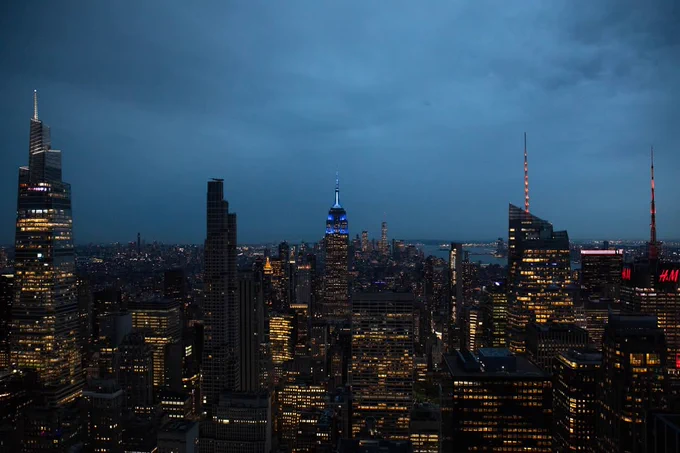A panoramic view of the New York City skyline at dusk, featuring the illuminated Empire State Building in the center with its blue lights. Surrounding skyscrapers are lit, and the sky is a deep blue.