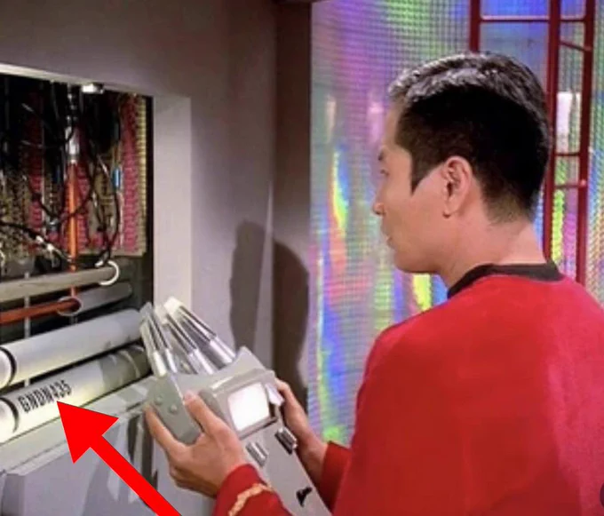 George Takei als Sulu in roter Uniform steht im Profil vor einem offenen Technikpanel der Enterprise und hält ein silbernes Gerät. Ein großer roter Pfeil zeigt auf das Label „GNDN435“ auf einem grauen Rohr
