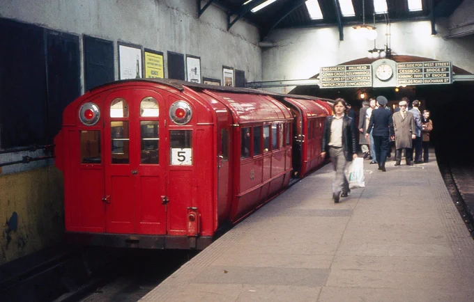 A bright red 1896 vintage Glasgow Subway train calls at Copland Road station (now Ibrox) on the outer rail service in April 1975. Its diminutive size compared to people on the very narrow island platform is clear. Above the tunnel mouths at the far end of the platform is a static display indicating which stations are reached from which platform, either side of a large clock.