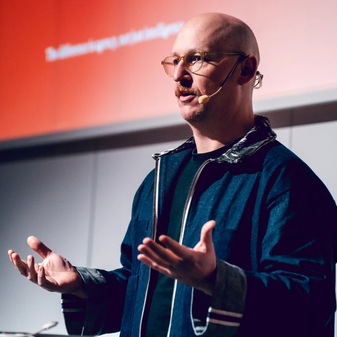 Speaker at Øredev in a dark jacket gesturing urgently with open hands against a red and gray backdrop.