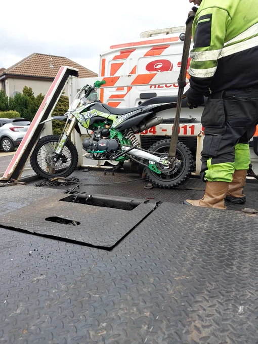 A green and black dirt bike is loaded onto a recovery truck by a person in high-visibility clothing.