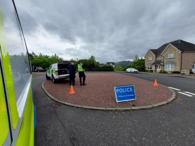 Police officers and vehicles at a scene with a 'Police Drone Operations' sign and orange cones.