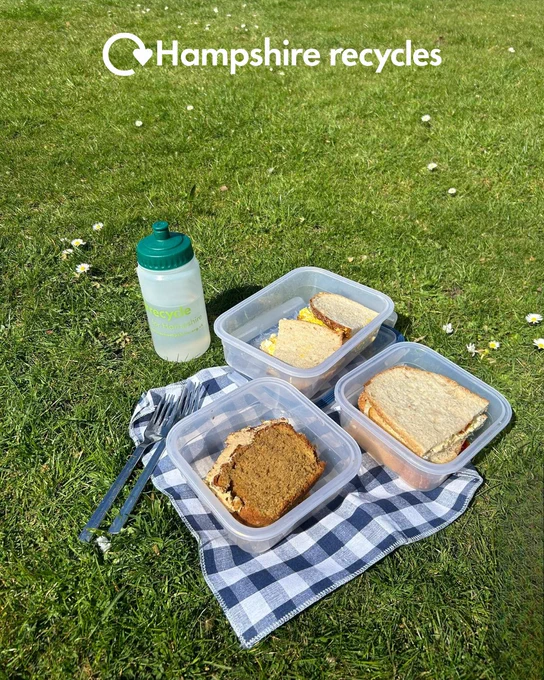 Photo of a picnic scene on grass with sandwiches and a slice of cake in reusable containers, reusable cutlery and a reusable water bottle on a blue gingham cloth. White Hampshire recycles logo in top centre.