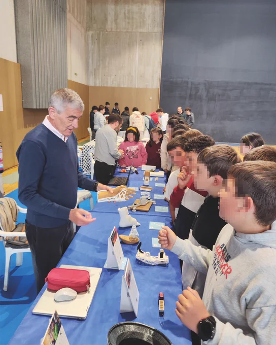 Voluntario de 3DLAN explica productos de apoyo impresos en 3D a un grupo de estudiantes en un stand con mesa azul