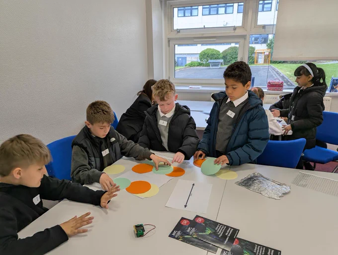 A group of young students is engaged in an educational activity at a table with colored paper circles. They appear focused and collaborative. In the background, two other students are working together at a separate table. The room is bright with a view of the outdoors through a window. Flyers are on the table.
