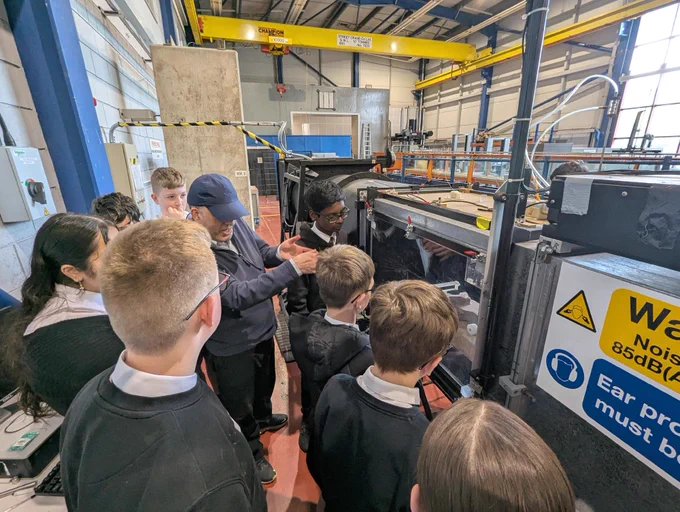 A group of people, some wearing uniforms, are gathered around a large piece of industrial equipment in a workshop setting. A person appears to be explaining the equipment operation. A warning sign indicates noise levels. The environment features high ceilings and metal structures.