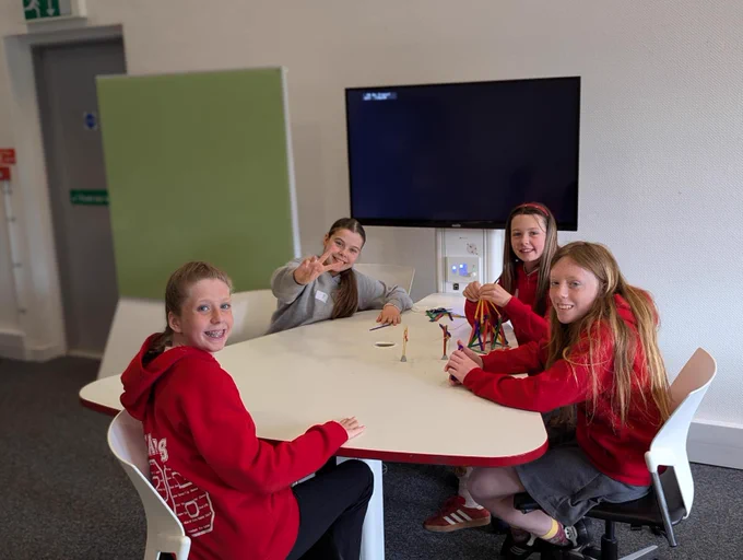 Four young students sit around a table in a classroom, engaging in an activity with colorful sticks. A large screen is mounted on the wall behind them. They appear to be enjoying their time together.