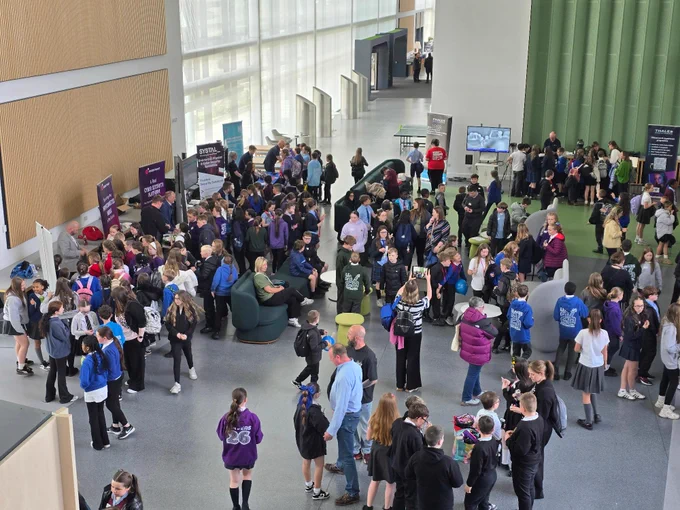 A large group of people gathered in a spacious indoor area with high ceilings and large windows. Some are standing near informational booths with signage, while others are seated in small groups. The environment appears busy and social.