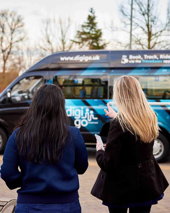 Two women look at a minibus with 'digigo.uk' and 'digi go' branding. One woman holds a phone.