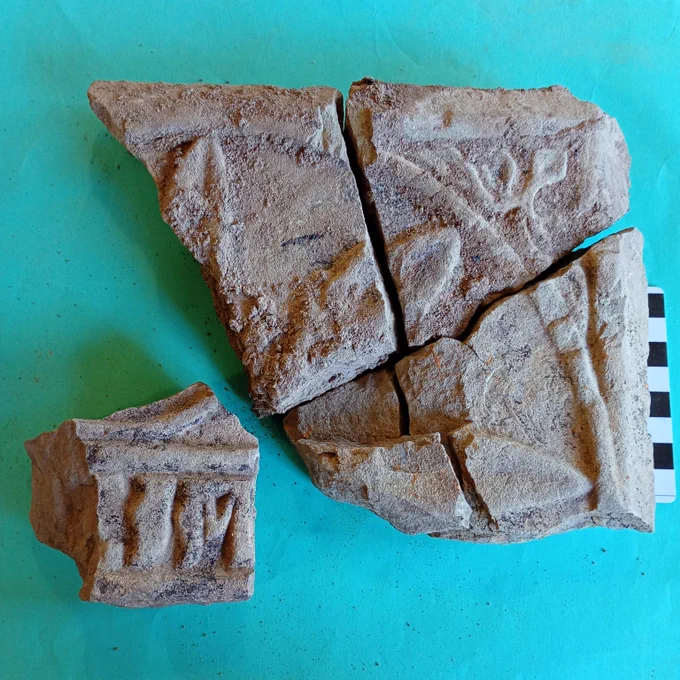 A piece of grey carved stone broken into four pieces. The stone is carved with a pattern of leaves. A fifth piece is carved with letters, possibly an 'i' and a an 'm'.