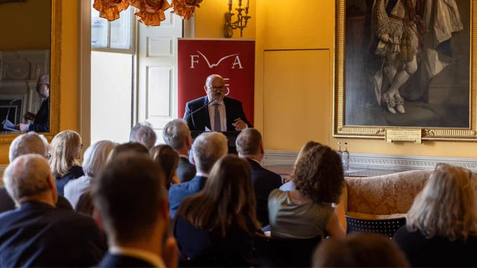 Belgian Interior Minister Bernard Quintin is speaking at a podium in a formal room, with a red banner displaying "FWA" behind him. The audience is seated, facing the speaker. A large painting is on the wall to the right. The atmosphere is professional and attentive.
