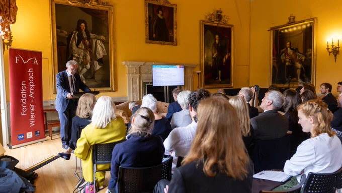 FWA President Professor Didier Viviers stands at a podium, giving a presentation to an audience in a room adorned with large, historic paintings. A Fondation Wiener-Anspach banner is visible beside the speaker. A screen displays content related to the presentation. The setting is elegant, with yellow walls and ornate decor.