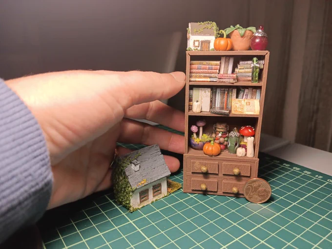 Fotografía de una maqueta en miniatura: una estantería con libros, pequeños objetos decorativos y tres cajones. Encima hay figuras diminutas como un mini invernadero, una calabaza y otros elementos. A la izquierda, una mano sostiene una pequeña casa en miniatura. En la base se ve una moneda de 2 céntimos que indica la escala.