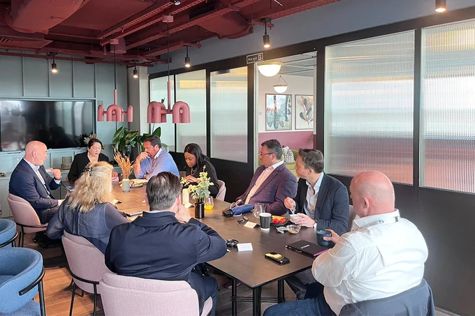 A group of people in a meeting room gather around a rectangular table. Some are engaged in conversation while others listen. The room has modern decor with a large screen on the wall and plants as decoration.