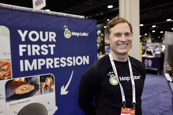 A smiling representative stands at a trade show booth for MapLabs. The booth features a blue backdrop with text and images promoting the brand.