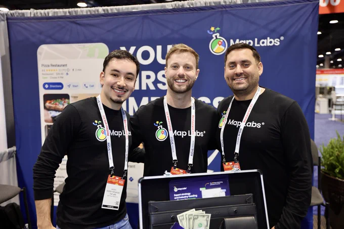 Three men in branded shirts smile at a trade show booth with a Map Labs backdrop. The scene conveys a professional and friendly atmosphere.