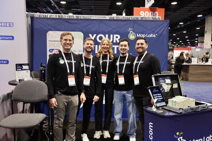 A group of five people at a trade show booth for Map Labs, smiling and wearing branded shirts. The atmosphere is professional and friendly.