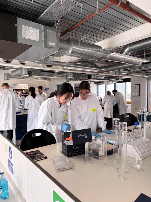 A group of people wearing lab coats work together in a modern laboratory. They are surrounded by scientific equipment and supplies, such as test tubes and beakers. The lab has visible ventilation ducts and a well-lit atmosphere.