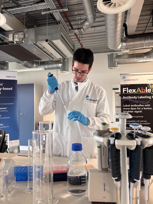 A person in a lab coat and gloves uses a pipette in a laboratory setting. Various lab equipment and containers are on the table. A banner for "Proteintech" and "FlexAble Antibody Labeling Kits" is visible in the background.