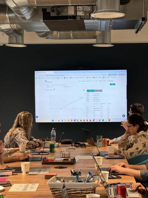 A group of people are seated around a conference table with laptops, attentively looking at a large screen on the wall displaying a graph and data from a website. The room has exposed ductwork in the ceiling and a dark green accent wall.