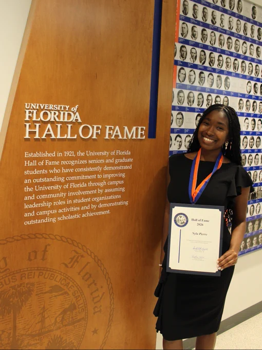 Nyla Pierre stands smiling beside the University of Florida Hall of Fame display while holding her Hall of Fame certificate.