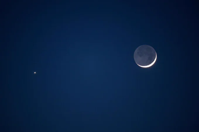 La Luna en fase creciente y Venus brillan en el cielo nocturno durante una conjunción. La Luna aparece en la parte superior derecha, con una delgada franja iluminada y el resto de su superficie tenuemente visible. Venus se ve como un punto brillante debajo y a la izquierda de la Luna. En la parte inferior se distingue la parte superior del Monumento a Washington iluminado, tras tejados oscuros de edificios.