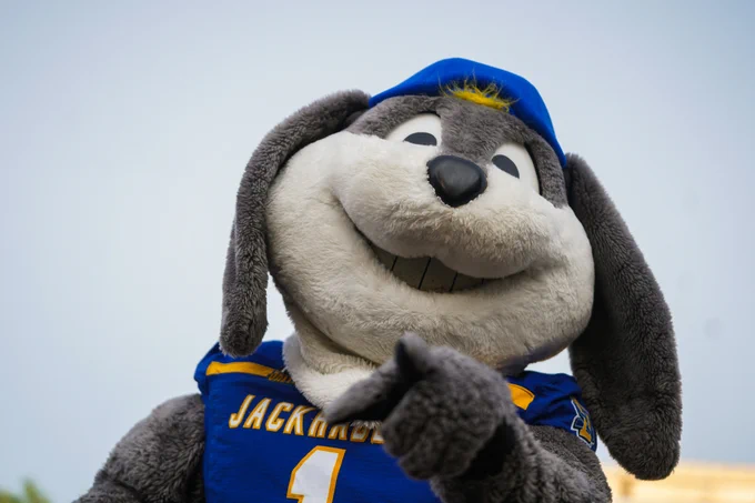 The SDSU Jackrabbits mascot is shown in a close‑up, low‑angle view wearing a blue jersey with “Jackrabbits” and the number 1, along with a blue cap, pointing toward the camera with one hand. The background is a plain, overcast sky, with no other visible elements in the frame.