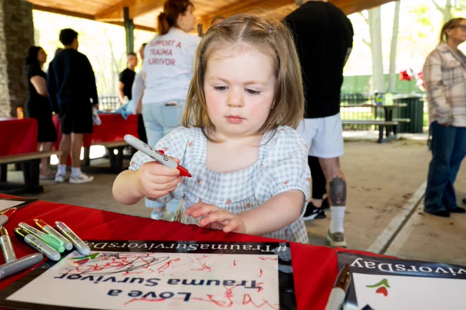 A child is focused on drawing with red marker on paper at an event. The surroundings include tables with markers and papers, other people interacting, and a T-shirt with "Trumpet Trauma Survivor" printed on it. The scene is in a covered outdoor area.