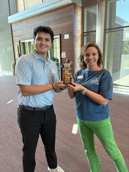 Two people stand indoors smiling while holding a small trophy with a goat figure on top. The person on the left wears a light blue collared shirt with dark pants, and the person on the right wears a blue “Surry-Yadkin Works” shirt with bright green pants. They are standing in a large, well-lit lobby with tall windows and wood-paneled walls.