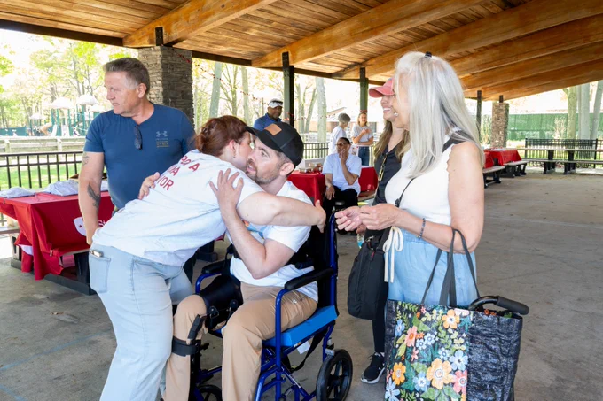 A group of people gathered under a pavilion, one sitting in a wheelchair receiving a hug. Others stand around them, some smiling. There are red table covers in the background.