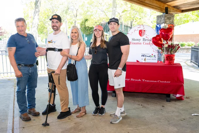 A group of five people stands together under a pavilion. One person uses a forearm crutch and is assisted by another individual. A table nearby is covered with a red cloth that reads "Stony Brook Trauma Center" and has a display of balloons. There are brochures and promotional items on the table. Trees and a fence are visible in the background.