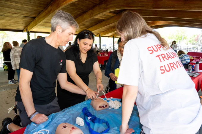 A group of people under a wooden pavilion are gathered around a table practicing medical procedures on trauma mannequins. One person wears a shirt with the text "Support Trauma Survivors.