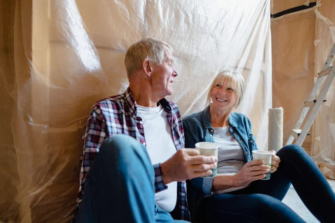 Elderly couple sitting on the floor with plastic-covered walls, holding cups and taking a break from home renovation.