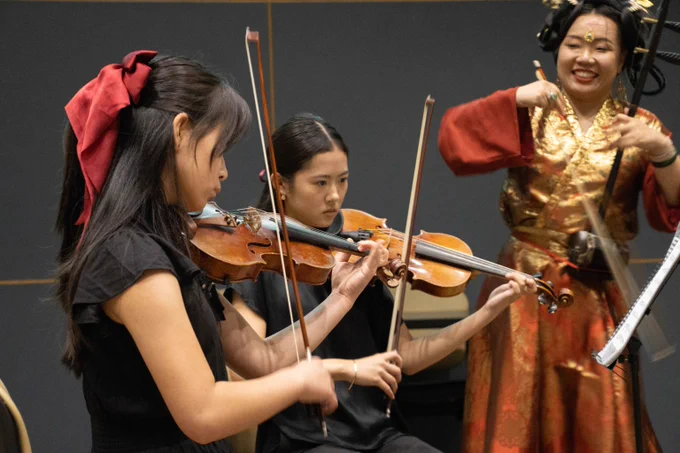 Two young violinists playing their instruments during a performance with an erhu performer in traditional attire playing along.