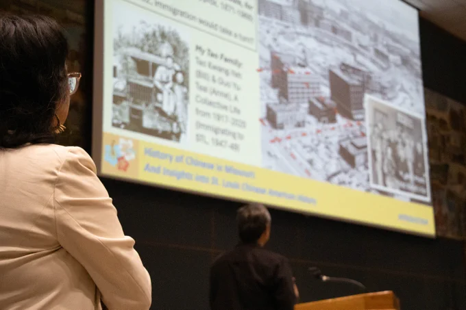 Director Anna Hui attentively watches a detailed historical presentation projected on a large screen in a lecture hall.