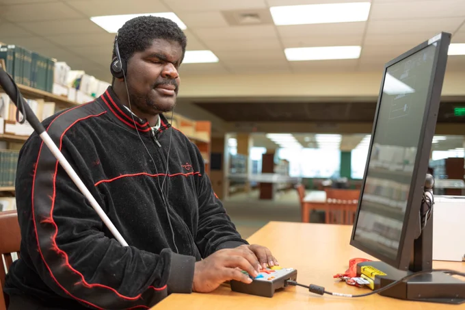 A young man sits at a desktop computer using a tactile device and wearing headphones, with a white cane beside him.