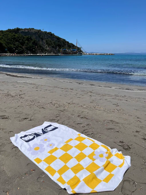 Açık, bulutsuz mavi bir gökyüzünün altında uzanan sakin ve berrak turkuaz renkli bir deniz manzarası. Arka planda koyu yeşil ağaçlarla kaplı dik bir tepe, küçük bir iskele ve denizde demirlemiş ahşap gövdeli, direkli bir gezi teknesi yer alıyor. Ön planda ise ıslak, kum rengi sahilin üzerine serilmiş beyaz renkli bir plaj havlusu bulunuyor. Havlunun üzerinde sol üst köşede lacivert renkli büyük harflerle "DIVA" yazısı ve hemen altında daha küçük puntolarla devam eden bir metin göze çarpıyor. Havlunun geri kalan bölümü ise sarı ve beyaz renklerden oluşan dama tahtası (kareli) deseniyle kaplı olup, bu desenin üzerinde açık mor ve beyaz renklerde iki adet büyük papatya motifi yer alıyor.