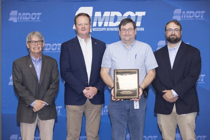A group of four people, including (from left to right) Craig Carter, Executive Director of ACEC/MS; Wayne Black, Garver Roadway Practice Leader; Billy Owen, MDOT District 6 Engineer; and Ross Baker, Assistant to Southern District Transportation Commissioner Charles Busby, stands posing for a photo in front of a blue backdrop with the "Mississippi Department of Transportation" logo. Billy Owen is holding a plaque.