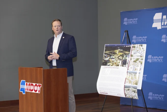 Wayne Black, Garver Roadway Practice Leader, stands at a podium with the MDOT logo, speaking at a presentation. A display board with images and text about the I-10 Diamondhead infrastructure project is positioned nearby. The backdrop features a step-and-repeat panel with MDOT branding.