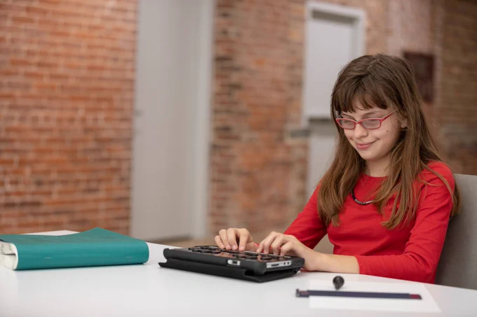 A girl types on a Braille device while sitting at a table.