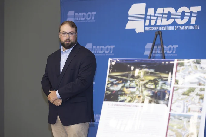 Ross Baker, Assistant to Southern District Transportation Commissioner Charles Busby, stands speaking at a presentation. A display board with images and text about the I-10 Diamondhead infrastructure project is positioned to the right. The backdrop features a step-and-repeat panel with MDOT branding.