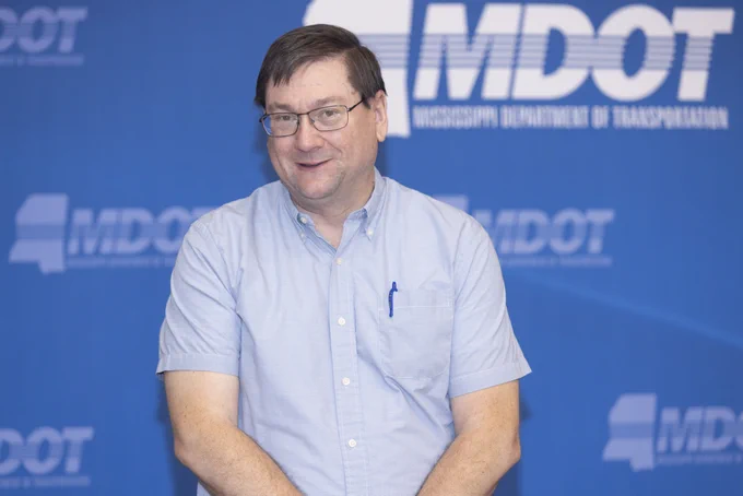 MDOT District 6 Engineer Billy Owen smiling in front of a blue backdrop with the "Mississippi Department of Transportation" logo and the abbreviation "MDOT" repeated. He is wearing a light blue button-up shirt with a pen in the pocket.